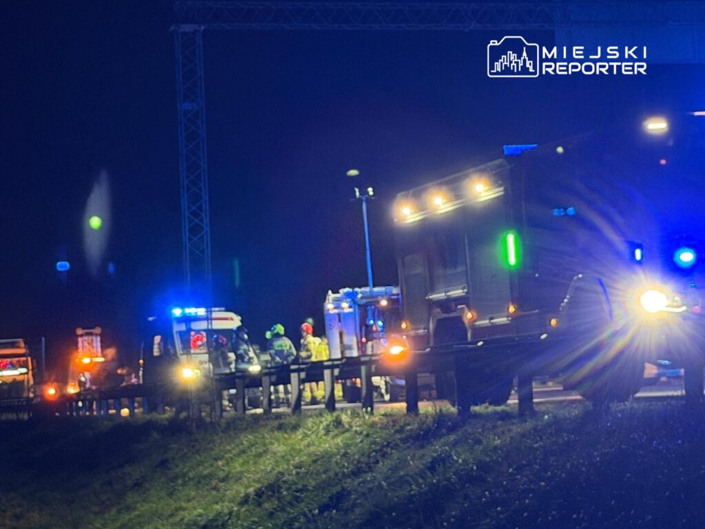 Grupa ratowników i strażaków pracuje przy wypadku drogowym, otoczona pojazdami z włączonymi sygnałami świetlnymi.