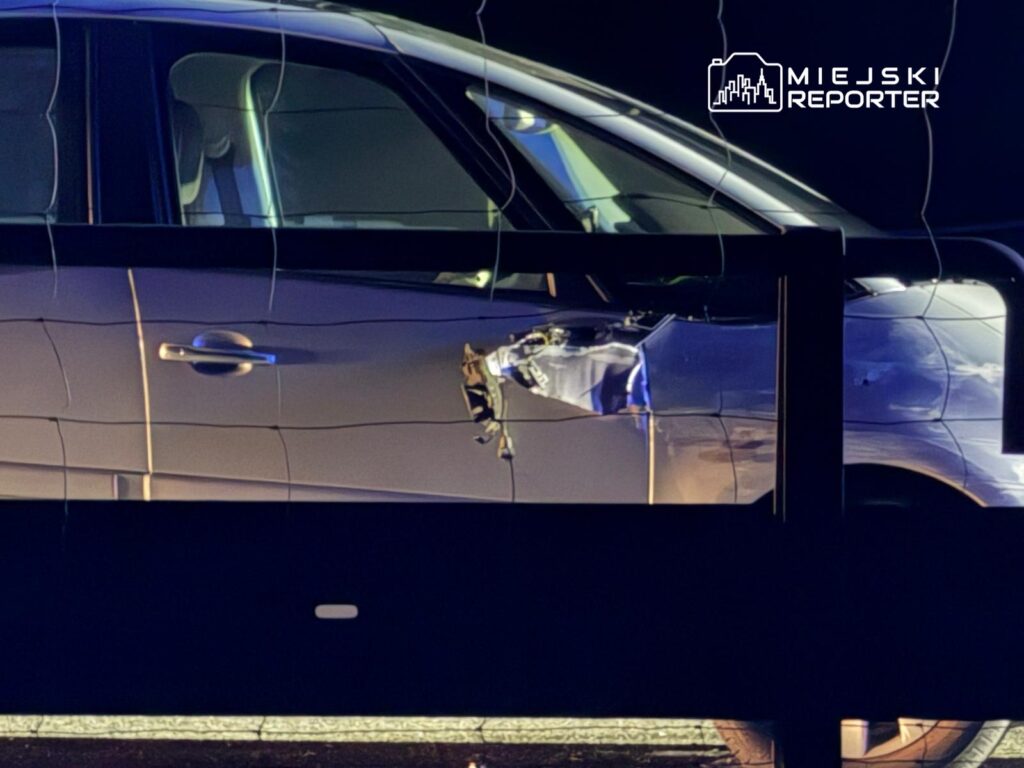 Uszkodzony samochód osobowy z wgniecioną boczną częścią, zaparkowany przy ciemnej barierce.