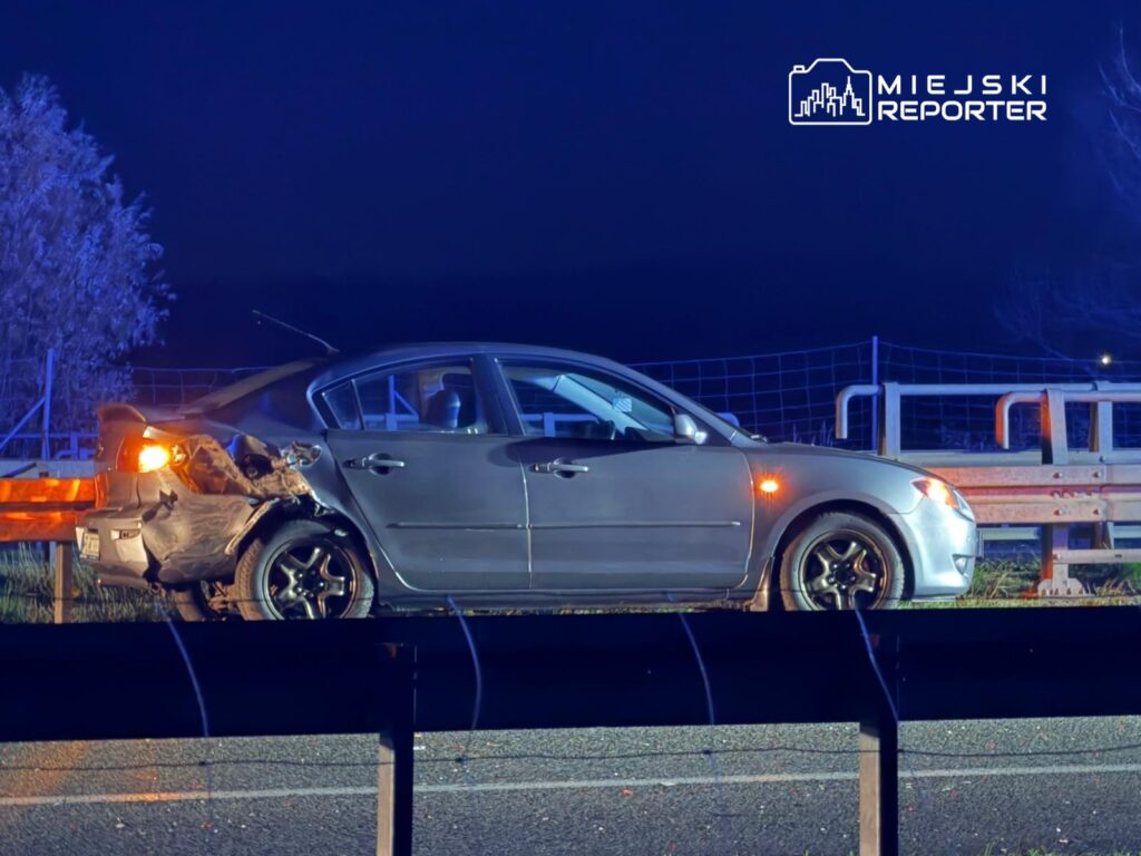 Uszkodzony samochód osobowy stoi na poboczu drogi w nocy, obok barierek ochronnych.