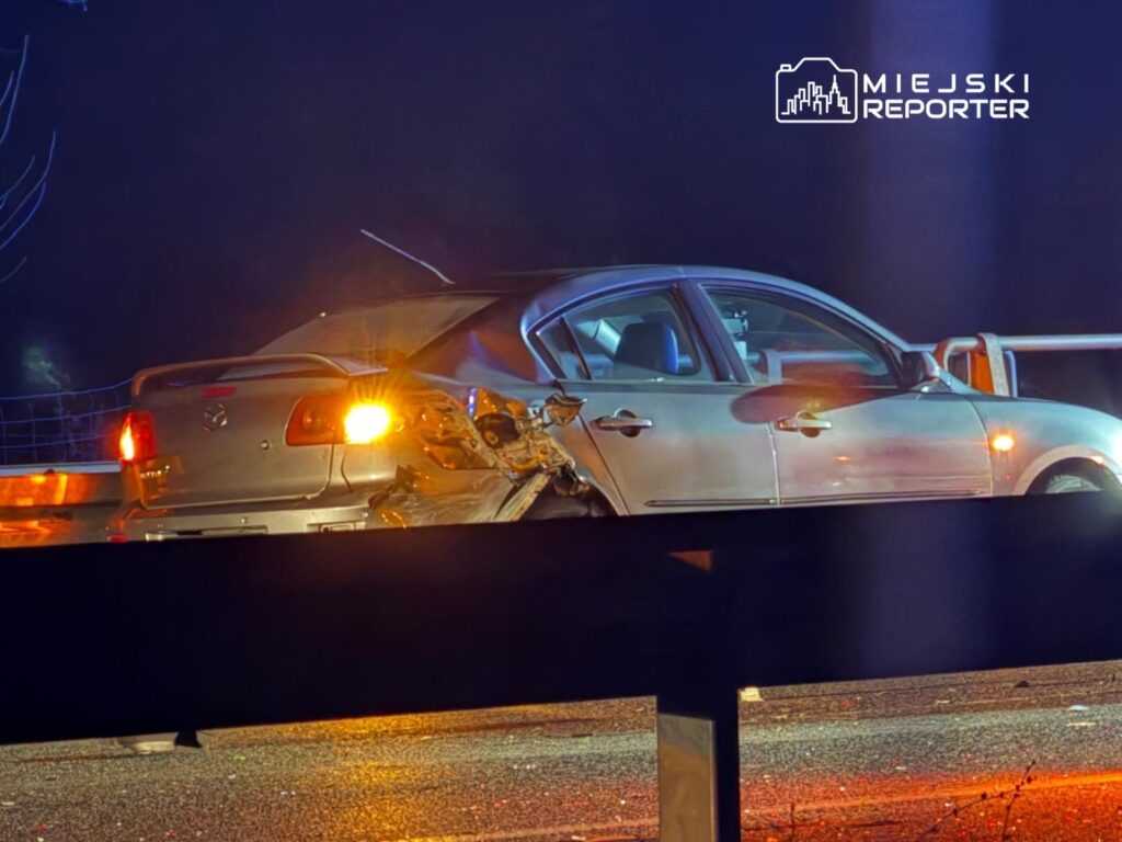 Uszkodzony samochód osobowy z wgniecioną tylną częścią stoi na poboczu drogi w nocy. W tle widoczne światła pojazdów.