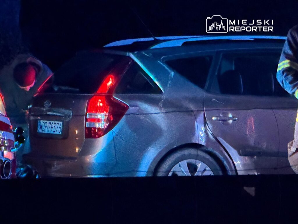 Mężczyzna w odblaskowej kurtce bada uszkodzone auto na drodze w nocy, przy zapalonych światłach innych pojazdów.