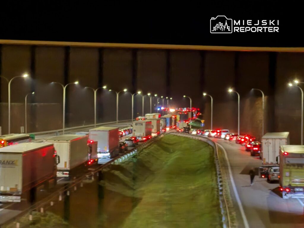 Długi korek samochodowy z ciężarówkami i osobówkami na drodze w nocy, oświetlony latarniami.