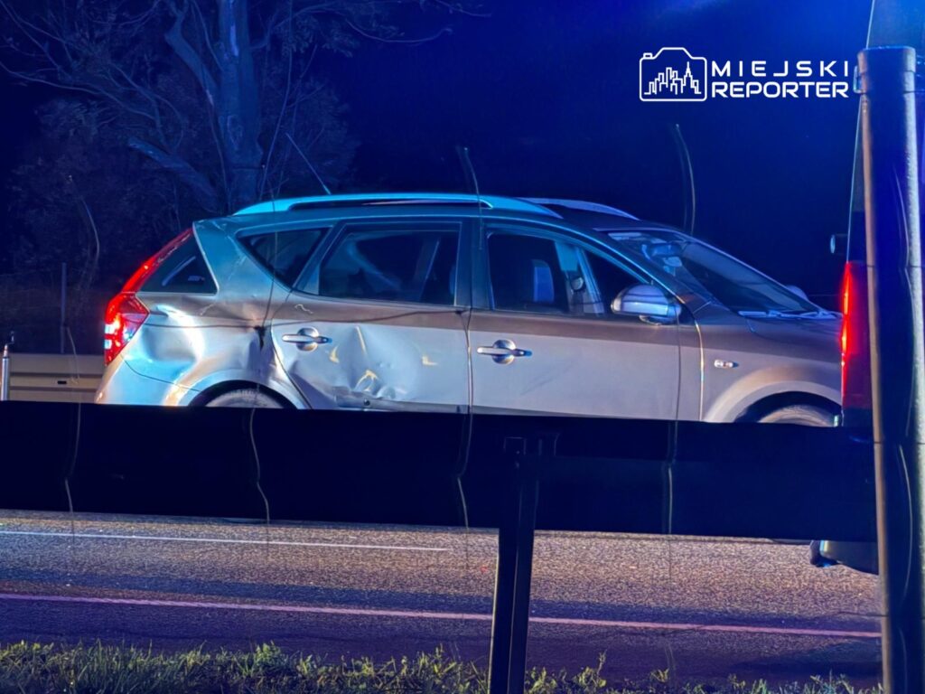 Uszkodzony srebrny samochód osobowy zatrzymany na poboczu drogi, w tle niebieskie światła policyjne.
