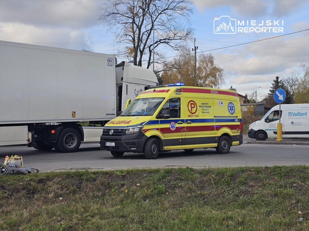 Karetka pogotowia z włączonymi sygnałami świetlnymi stoi obok dużego tira na drodze.