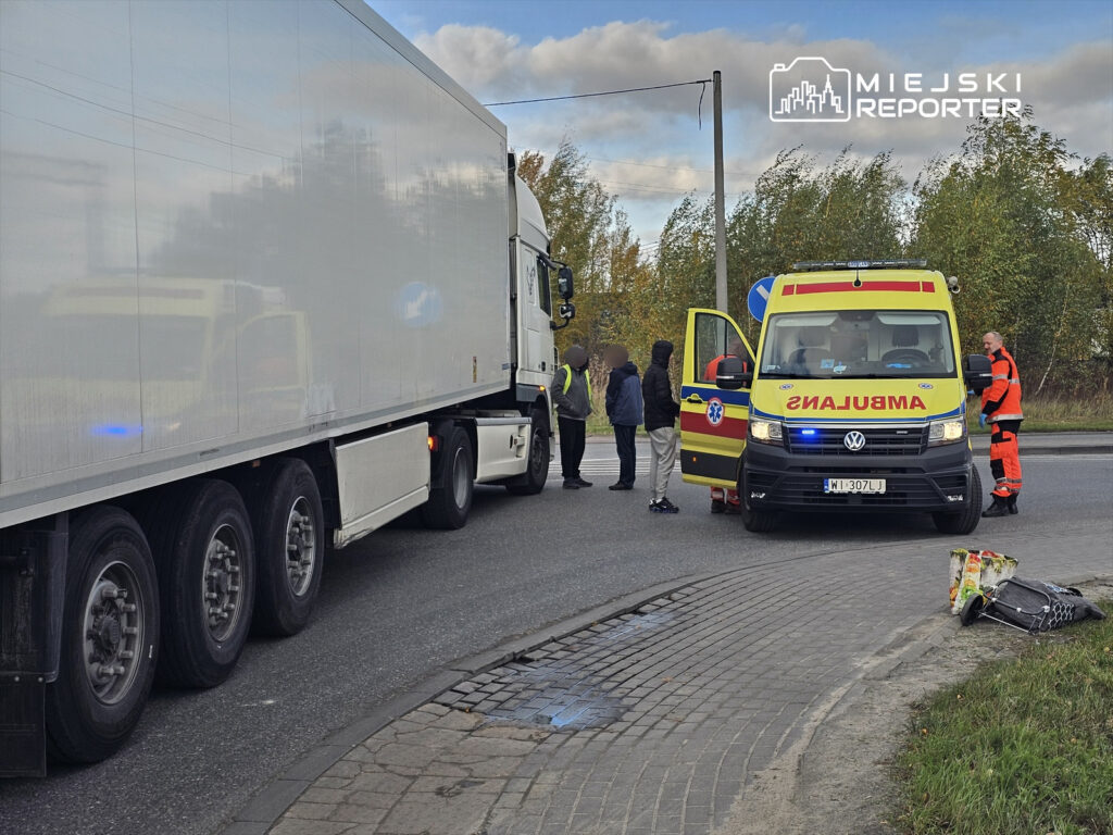 Karetka pogotowia zatrzymana przy drodze, obok stoją mężczyźni rozmawiający z ratownikami, w tle ciężarówka.