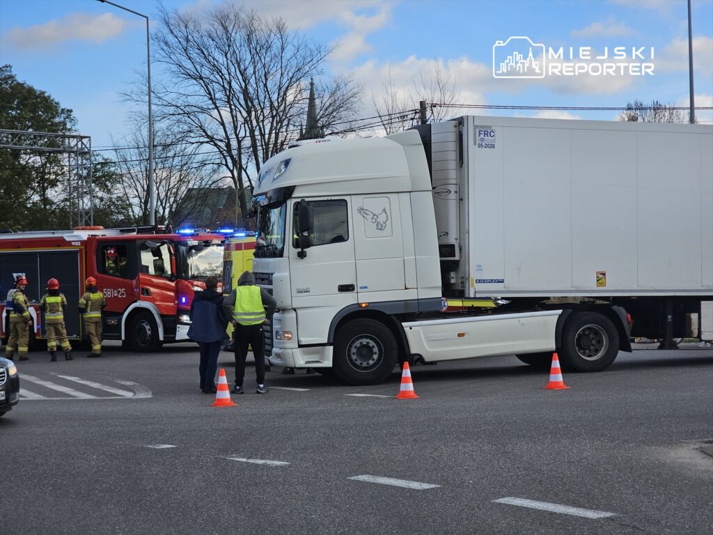 Czterech strażaków w mundurach rozmawia z mężczyzną w kamizelce odblaskowej obok ciężarówki, przy włączonym wozie strażackim.