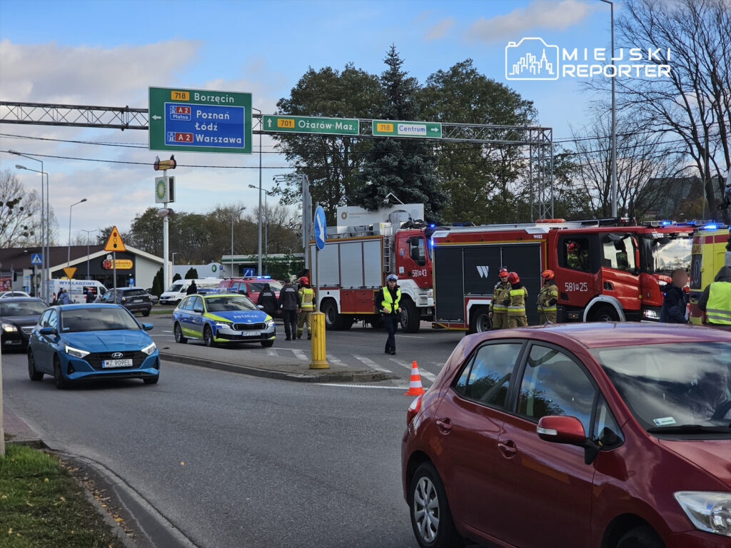 Na skrzyżowaniu ruchu drogowego stoją pojazdy ratunkowe, w tym wóz strażacki i radiowóz policji, a obok grupka strażaków.