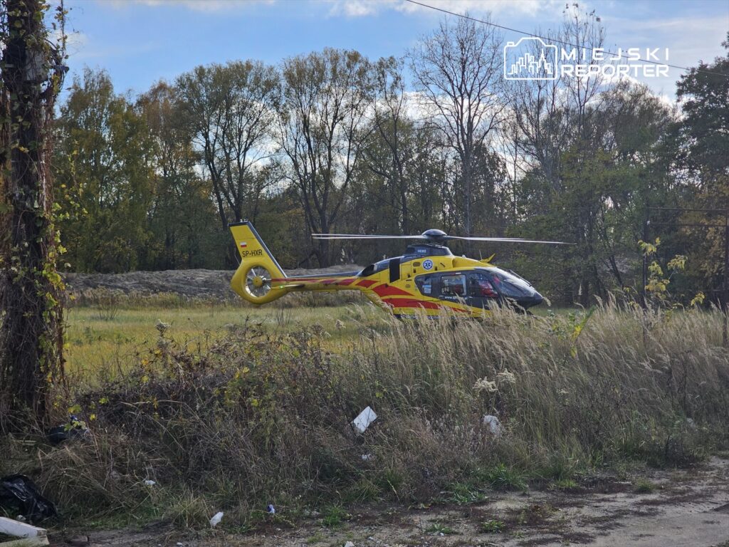 Helikopter ratunkowy ląduje na otwartym terenie, otoczony wysoką trawą i drzewami w tle.