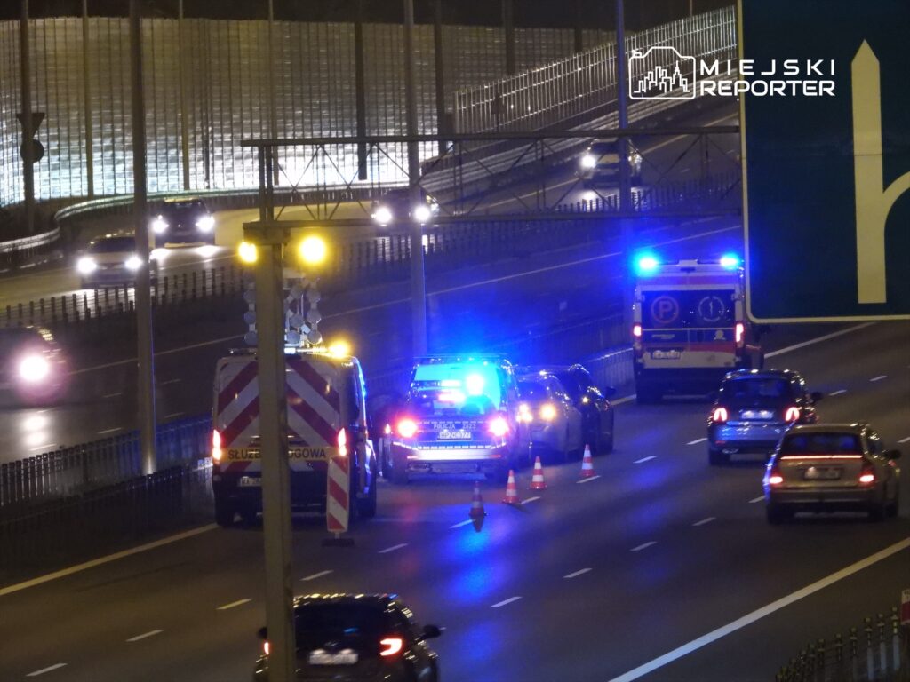 Na jezdni zatrzymane pojazdy służb ratunkowych z włączonymi sygnałami świetlnymi, otoczone przez trójkąty ostrzegawcze.