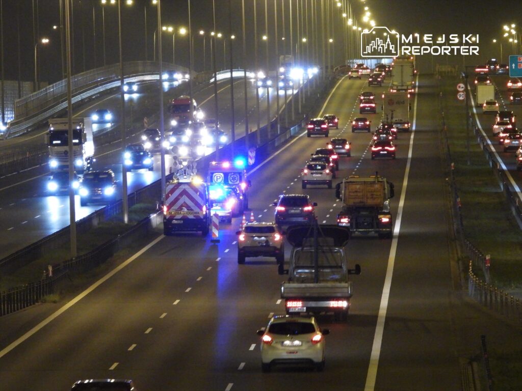 Na drodze ekspresowej nocą widoczny jest samochód z włączonymi sygnałami świetlnymi oraz kilka pojazdów osobowych.