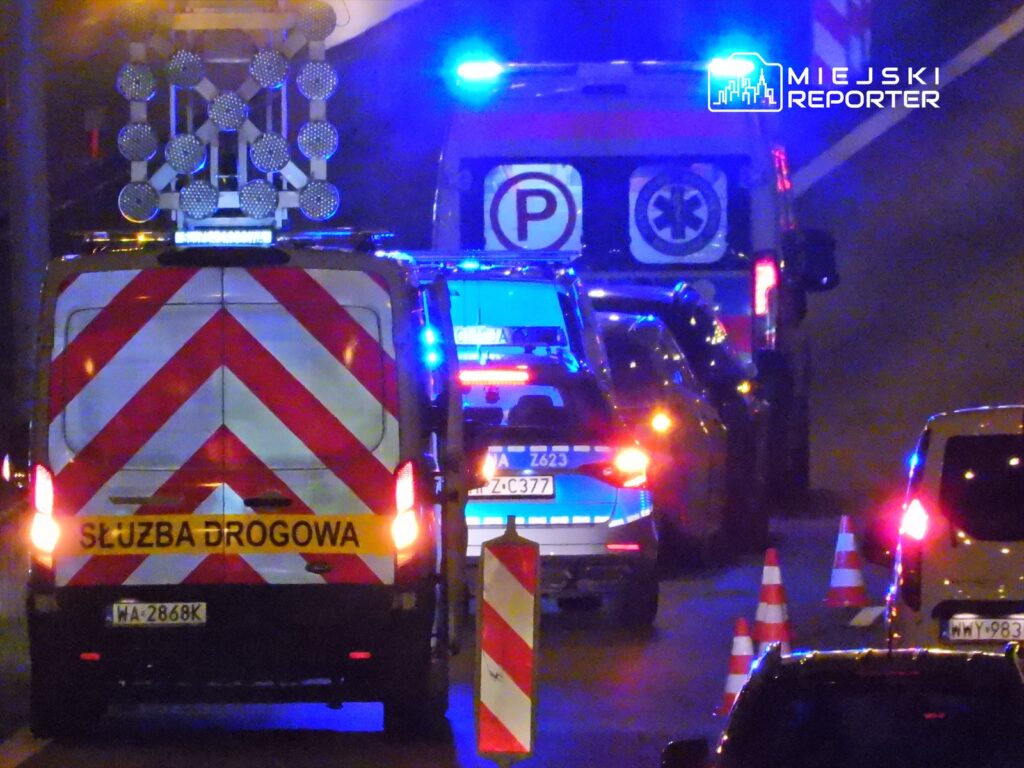 Na drodze zatrzymane pojazdy służb ratunkowych z włączonymi sygnałami świetlnymi, otoczone pachołkami.