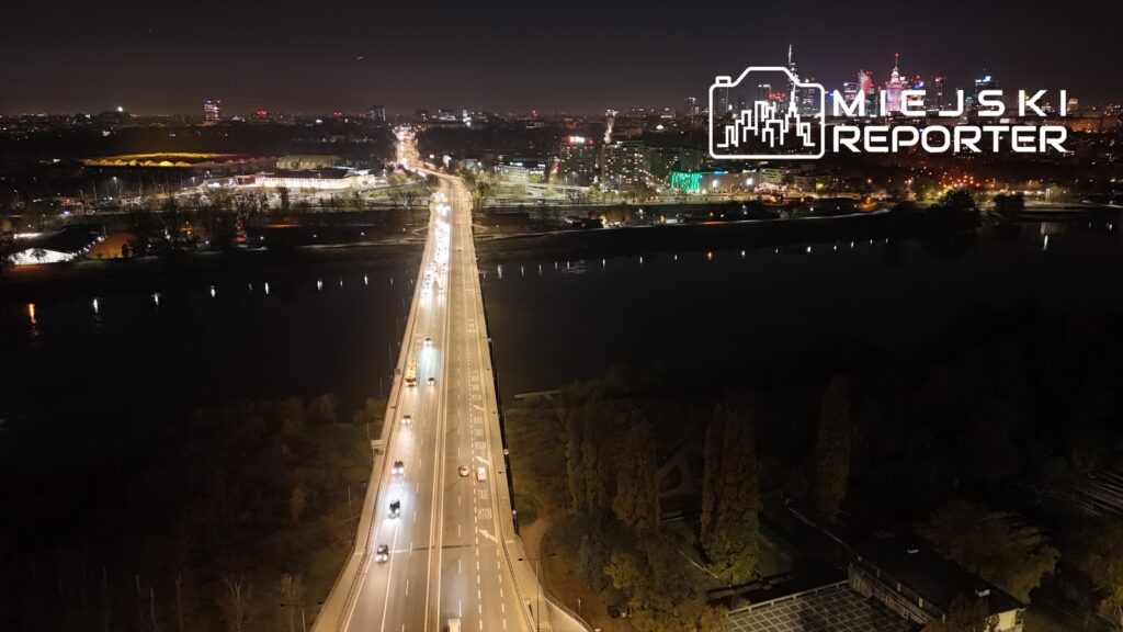 Widok na oświetloną drogę z ruchem samochodowym nad rzeką w nocy, z tłem miejskim w oddali.