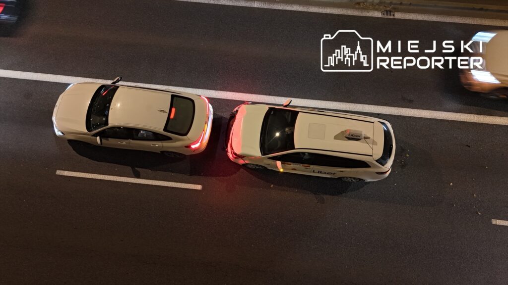 Dwa białe samochody osobowe, w tym jeden z logo Uber, stoją na jezdni w nocy, obok siebie.