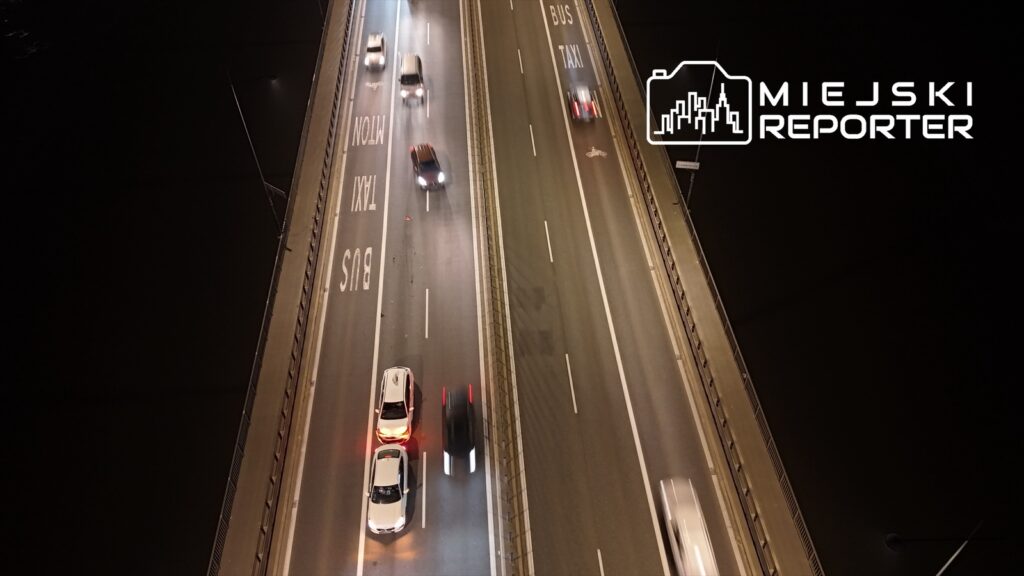 Ruch samochodów na oświetlonym moście z oznaczeniami drogowymi dla busów i taksówek.