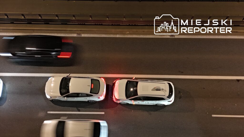 Dwa białe samochody osobowe z logo Ubera stoją obok siebie na ruchliwej ulicy nocą, wokół poruszają się inne pojazdy.