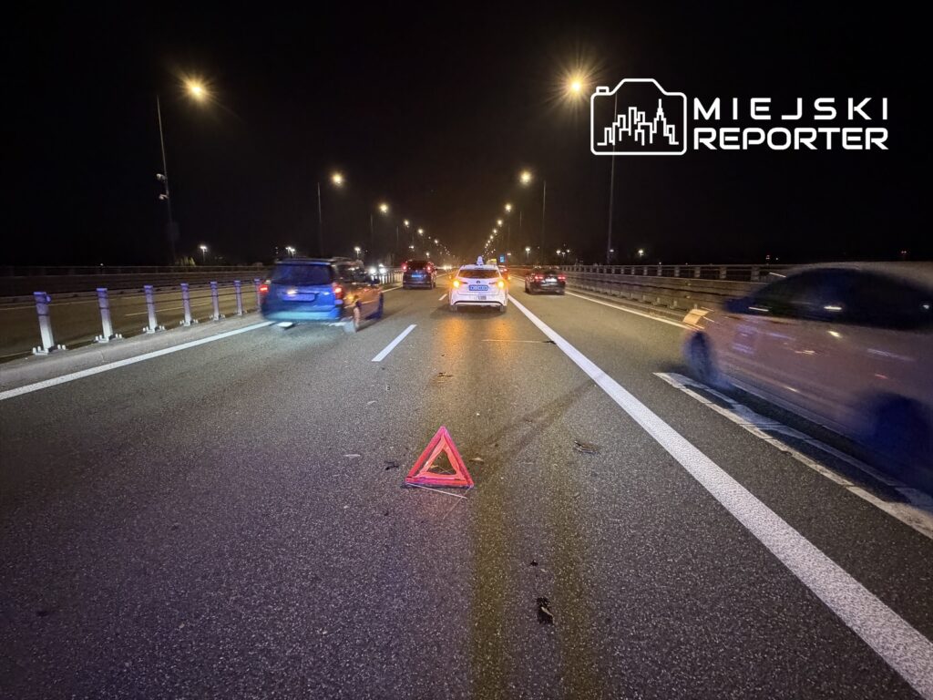 Czerwony trójkąt ostrzegawczy ustawiony na jezdni nocą, obok poruszające się samochody na drodze.