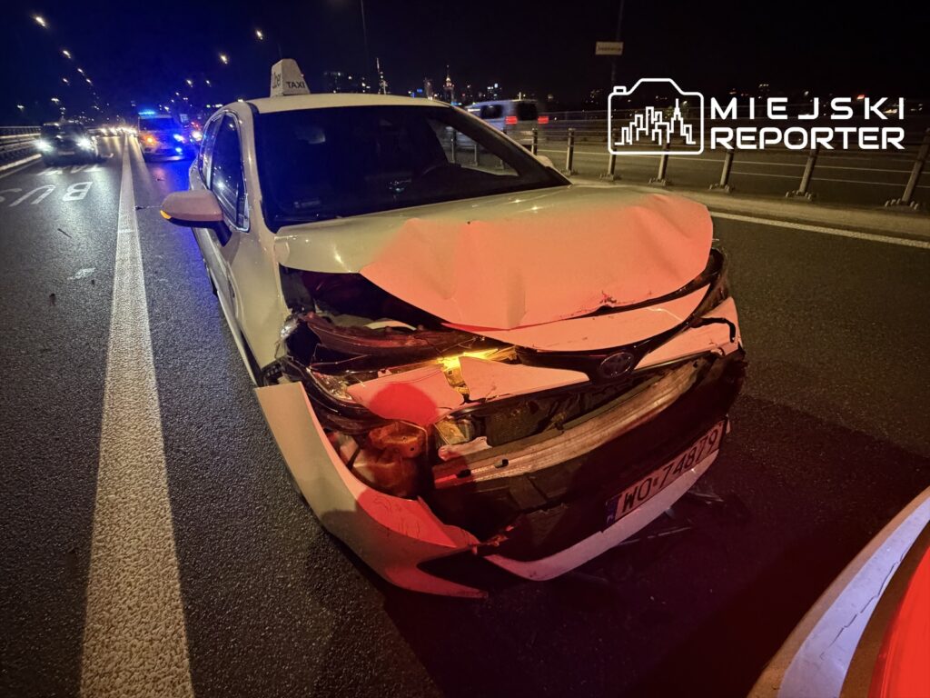 Uszkodzony samochód taxi z wgniecionym przodem stoi na jezdni, w tle widać policyjne światła.