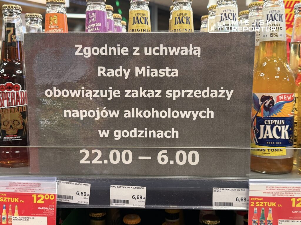 Tablica informacyjna w sklepie z napisem: "Zgodnie z uchwałą Rady Miasta obowiązuje zakaz sprzedaży napojów alkoholowych w godzinach 22.00 – 6.00". W tle widoczne butelki piwa.