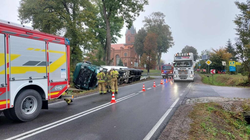 Strażacy pracują nad przewróconą ciężarówką przy drodze, obok nowoczesnego budynku z czerwonej cegły.