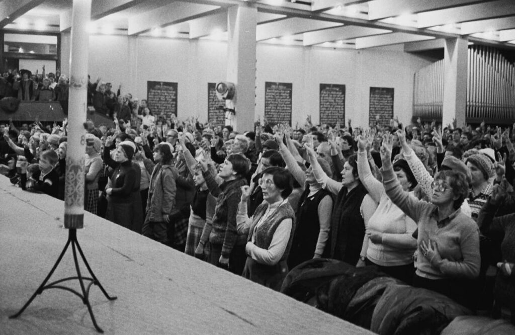 Tłum ludzi w podniosłych nastrojach podnosi ręce w geście aprobaty w dużej sali z oświetleniem sufitowym.