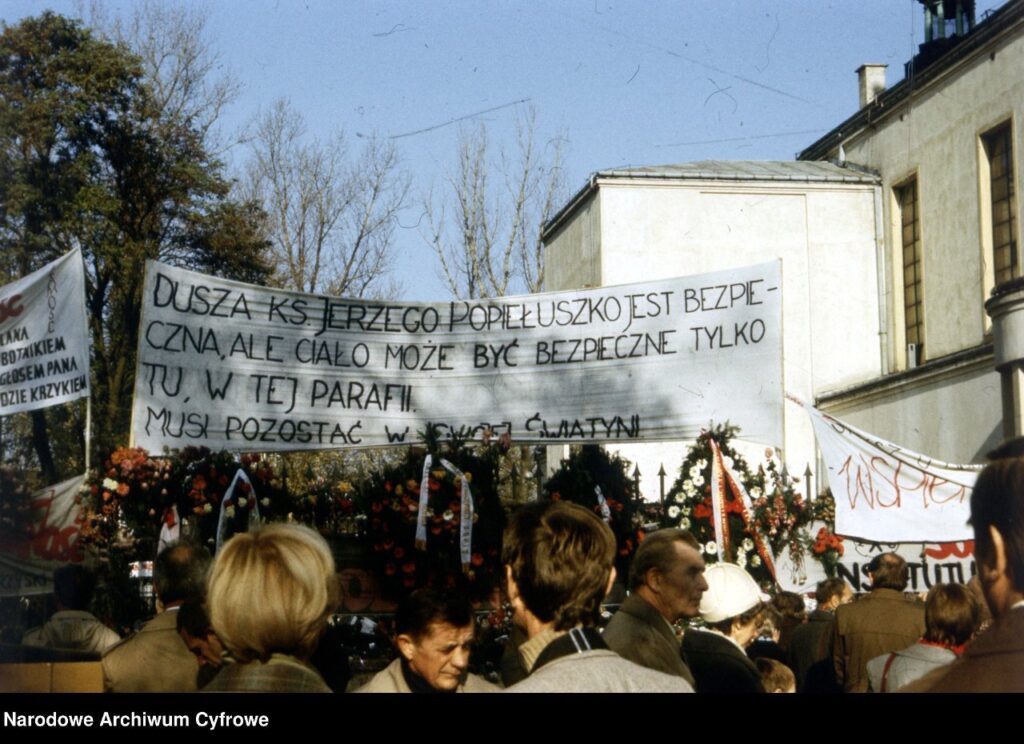 Ludzie z transparentami uczestniczą w zgromadzeniu przed budynkiem, w tle widoczne kwiaty i drzewa.