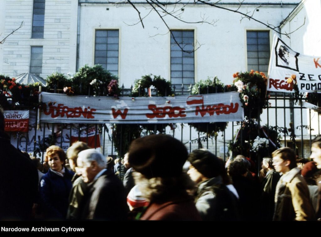 Tłum ludzi przechodzi obok bramy udekorowanej transparentem z napisem „Uderzenie w serce narodu” oraz kwiatami.