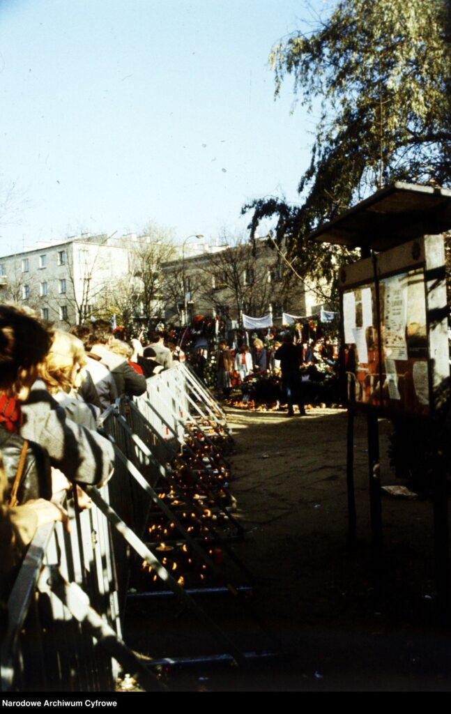 Ludzie stojący w kolejce przy ogrodzeniu, w tle widać kwiaty i znicze, a w powietrzu unosi się atmosfera zadumy.