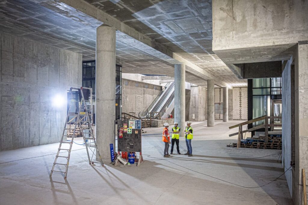 Grupa trzech pracowników budowlanych rozmawia w przestronnym wnętrzu w trakcie budowy, obok drabiny i sprzętu.