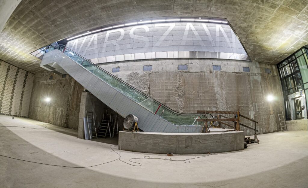 Wnętrze budynku z nowoczesną ruchomą schodami prowadzącymi do górnej części, z betonowymi ścianami i oświetleniem.