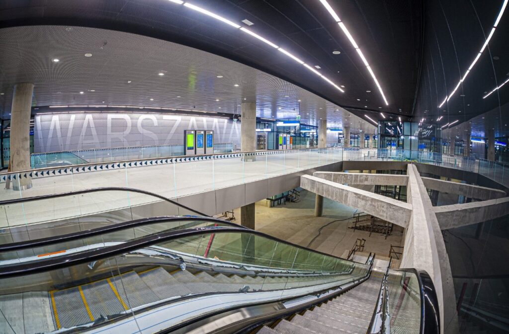 Nowoczesna stacja kolejowa z przeszkloną fasadą, na której widnieje napis "WARSZAWA", z ruchomymi schodami w tle.