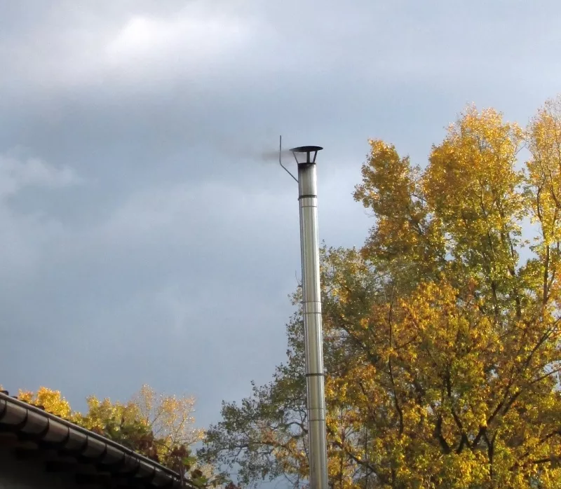 Dym wydobywający się z komina metalowego wśród drzew z żółtymi liśćmi pod szarym niebem.