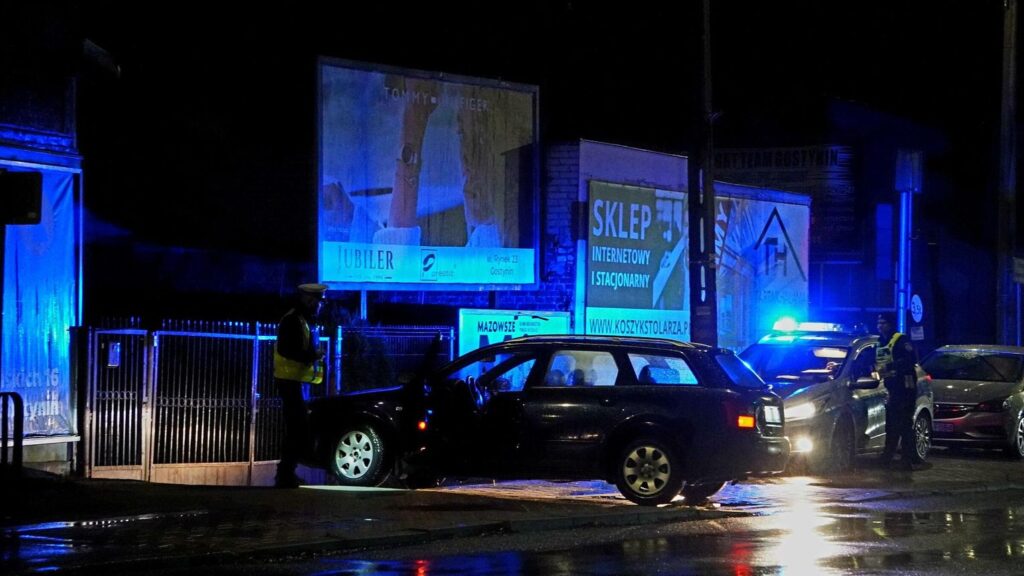 Policjant w odblaskowej kamizelce rozmawia z kierowcą w nocy przy zaparkowanym czarnym samochodzie.