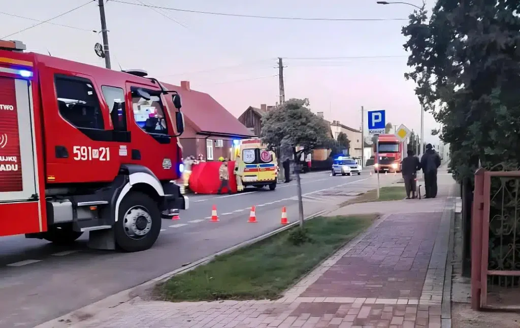 Czerwony wóz strażacki z włączonymi sygnalizatorami stoi na ulicy obok karetki, a ratownicy pracują przy poszkodowanym.