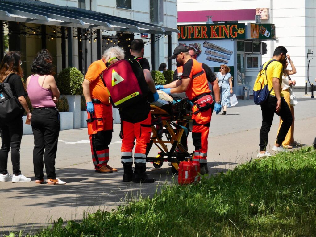 Zespół ratowników medycznych udziela pomocy osobie na noszach na ruchliwej ulicy. W tle przechodnie i lokalne sklepy.