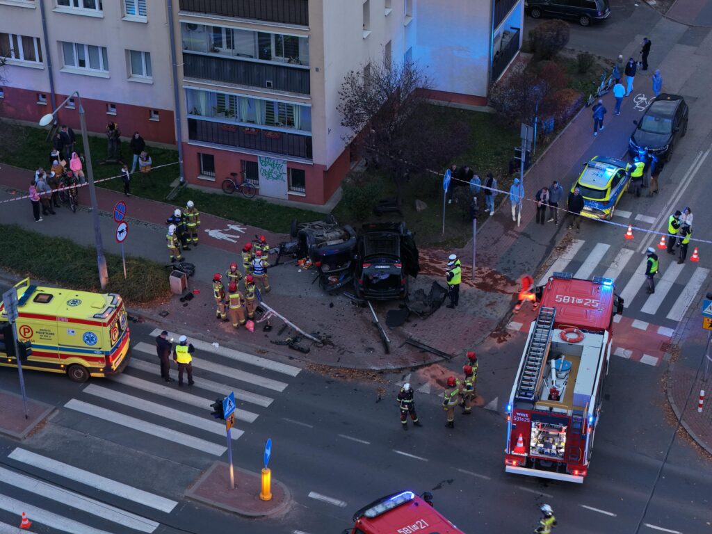 Grupa strażaków i ratowników medycznych pracuje przy przewróconym samochodzie na drodze, obok zgromadzonych gapiów.