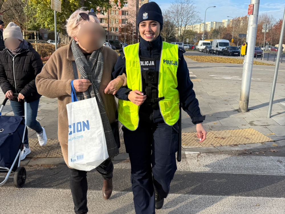 Policjantka w kamizelce odblaskowej asystuje starszej kobiecie z torbą, przechodząc przez chodnik w miejskim otoczeniu.