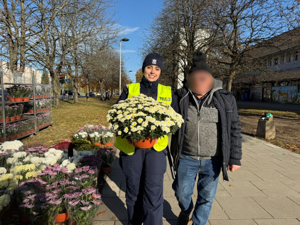 Policjantka w żółtej kamizelce trzyma bukiet białych kwiatów, stojąc obok mężczyzny na ulicy z drzewami w tle.