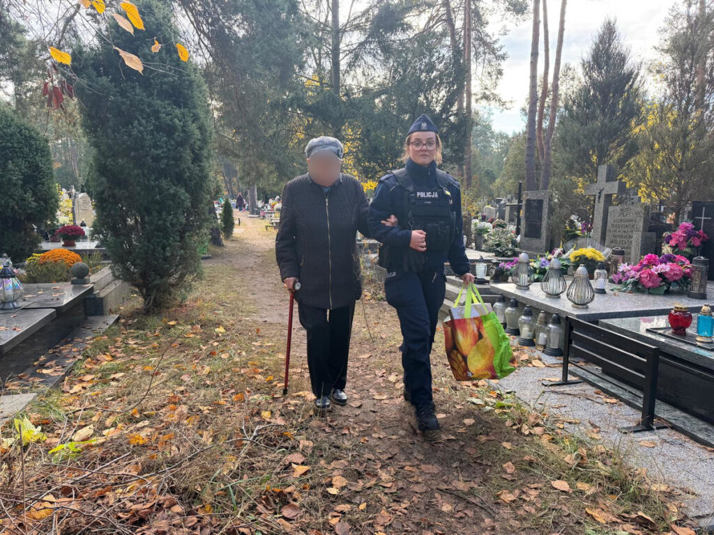 Starsza kobieta z laską idzie z policjantką, trzymającą torbę, na cmentarzu otoczonym grobami i kwiatami.