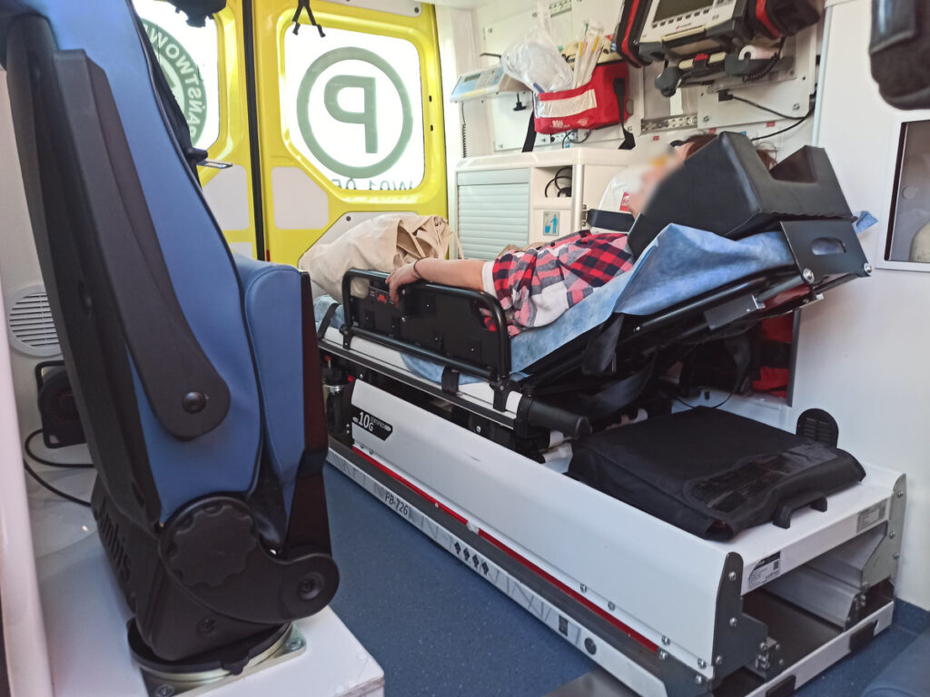 Kobieta leżąca na noszach w karetce, otoczona sprzętem medycznym i wyposażeniem ratunkowym.