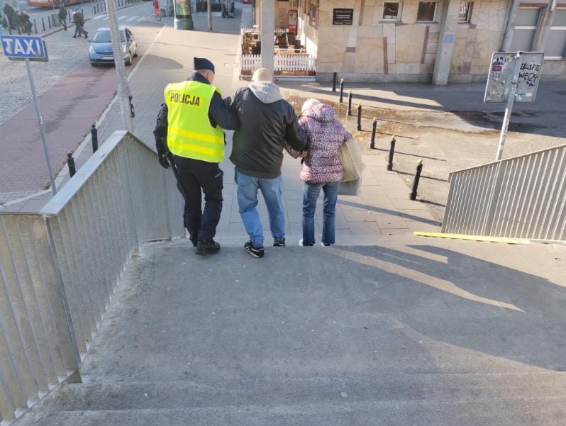 Policjant w żółtej kamizelce pomaga starszemu mężczyźnie i kobiecie schodzić po schodach w miejskim otoczeniu.