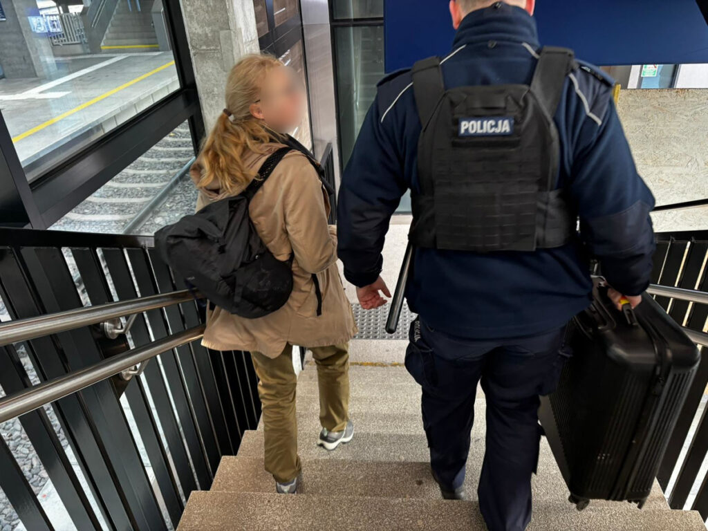 Kobieta z plecakiem schodzi po schodach, towarzyszy jej policjant niosący walizkę. W tle widoczne perony kolejowe.