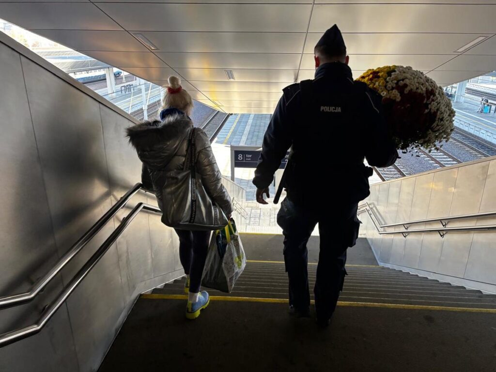 Kobieta w srebrnej kurtce schodzi po schodach, a obok niej policjant niosący bukiet kwiatów.