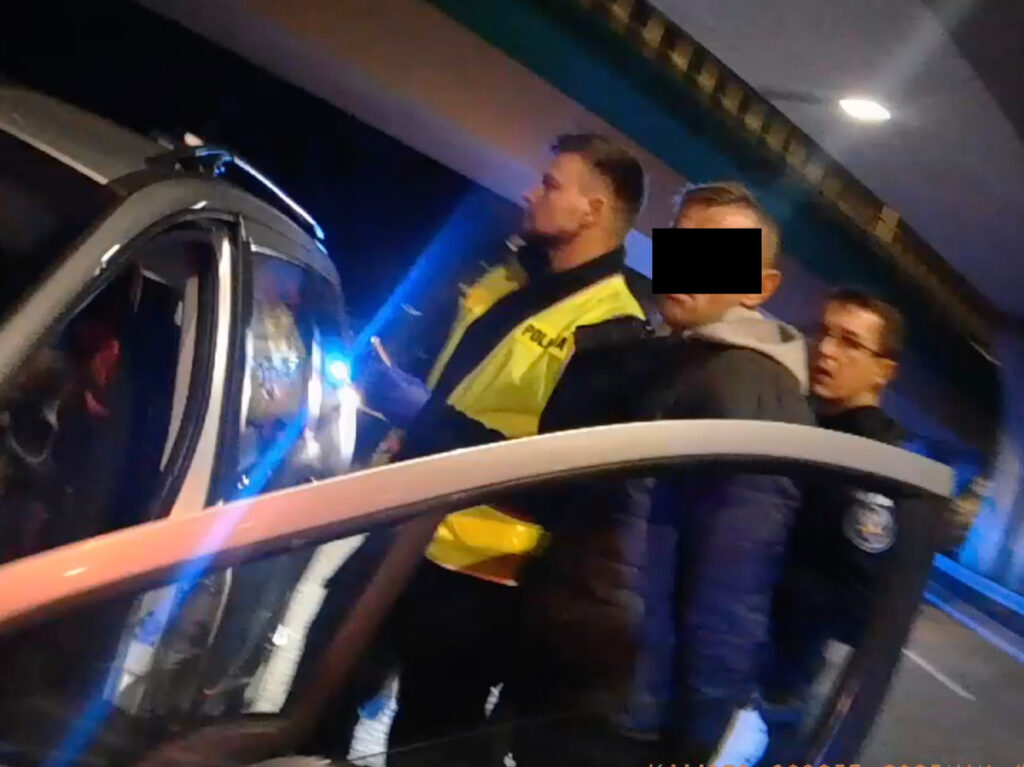 Mężczyzna w szarej bluzie jest zatrzymywany przez policjantów w odblaskowych kamizelkach w nocy.