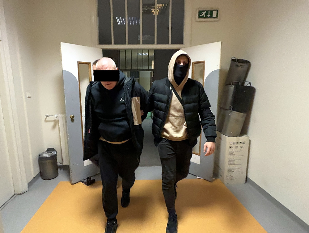 Mężczyzna w czarnej bluzie i dresach prowadzony przez drugiego mężczyznę w czarnej kurtce w korytarzu budynku.