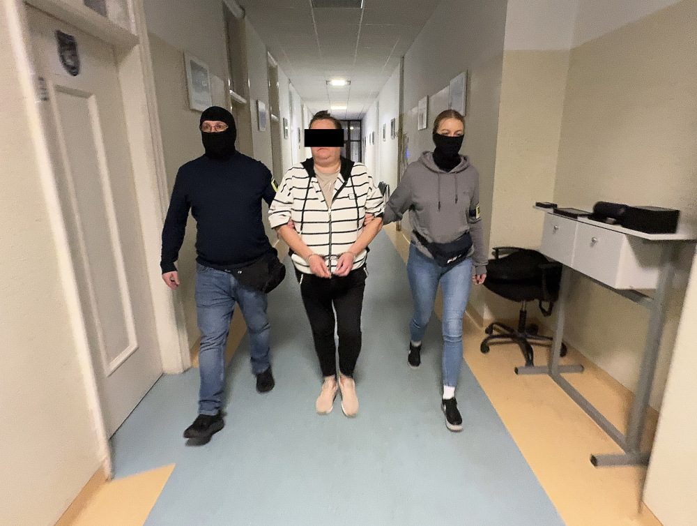 Trzech ludzi idących korytarzem w budynku, z jedną osobą w środku, trzymaną za rękę przez dwie osoby w maskach.