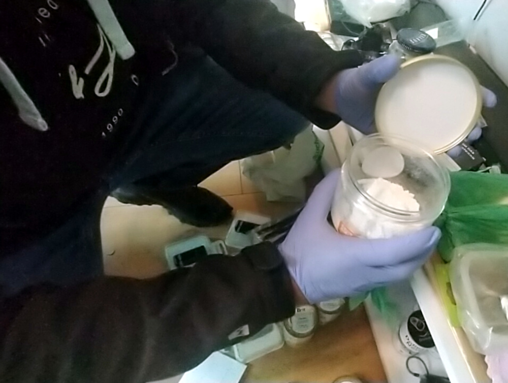 Mężczyzna w rękawiczkach otwiera słoik z białym proszkiem w zagraconym pomieszczeniu.