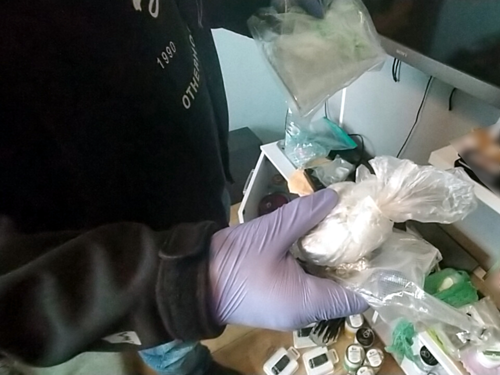 Mężczyzna w rękawiczkach lateksowych trzyma pakunek z białym proszkiem, stojąc obok rozłożonych przedmiotów na stole.