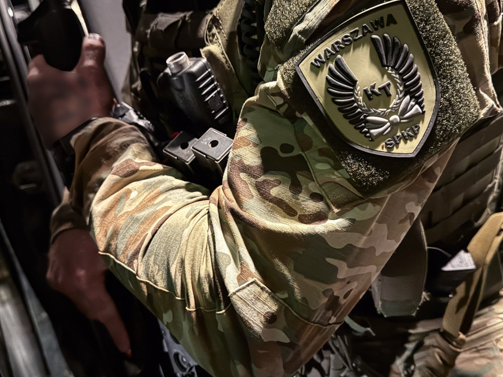 Żołnierz w mundurze kamuflażowym trzyma broń, a na ramieniu ma naszywkę z napisem "WARSZAWA KT".