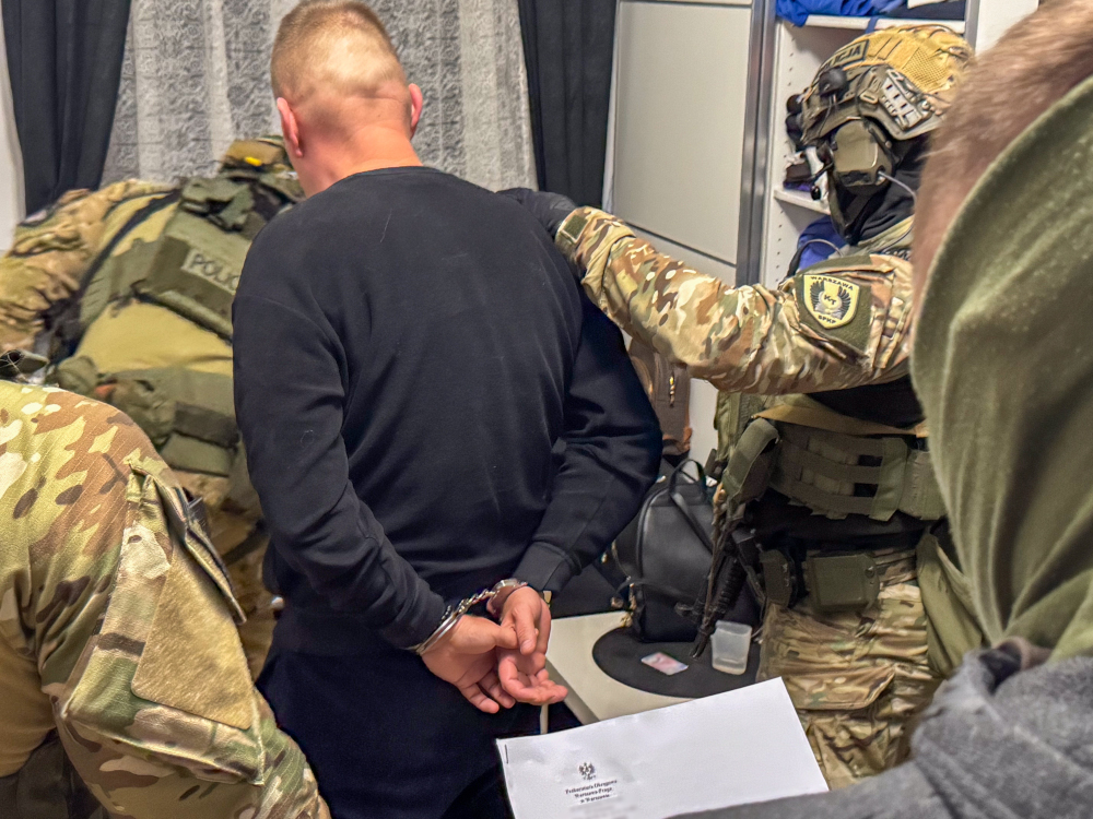 Mężczyzna w czarnej bluzie z rękami skuty w kajdankach, otoczony przez uzbrojonych funkcjonariuszy w kamizelkach.