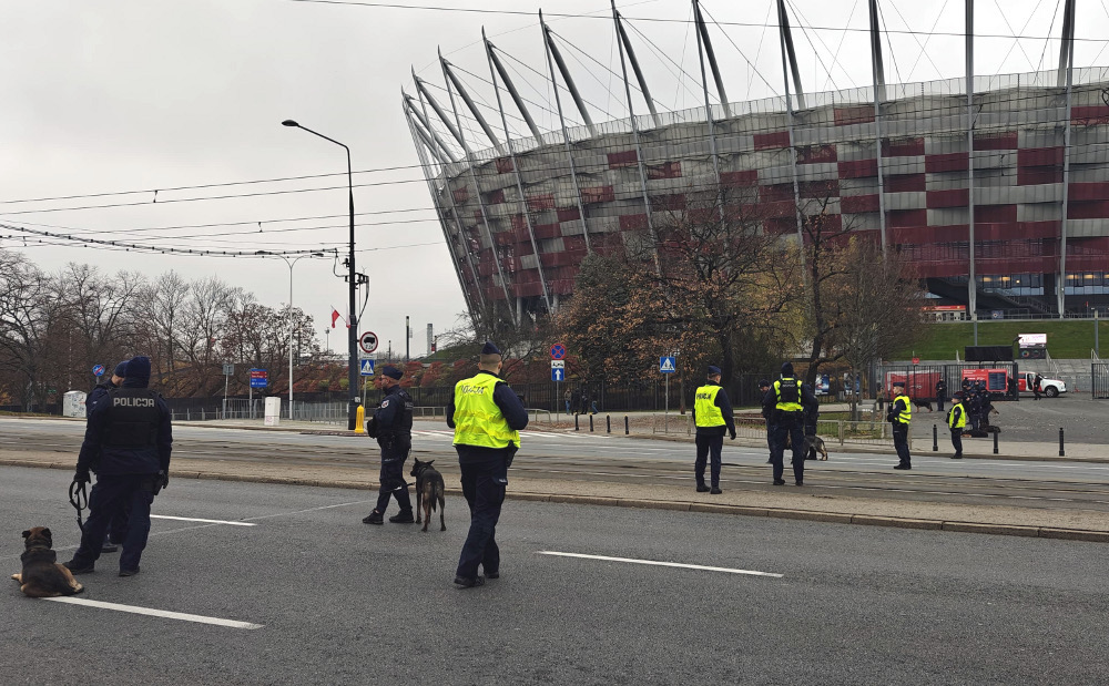 Grupa policjantów w kamizelkach odblaskowych prowadzi psy obok stadionu z charakterystyczną architekturą.